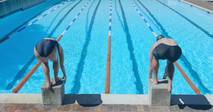 Swimmers wearing caps crouching on starting blocks in outdoor lap pool, with lane ropes and goggles