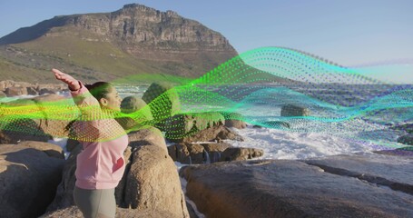 Standing Indian woman raising arms on coastal rocks, with ocean waves and digital ribbon overlay