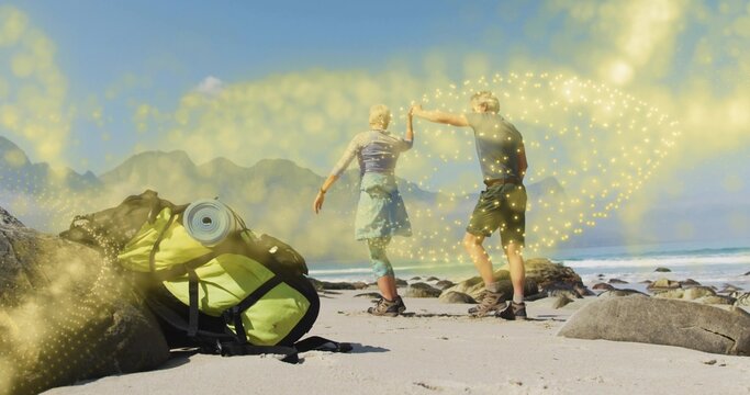 Hikers holding hands walking on beach near rocky outcrops wearing hiking boots, with green backpack