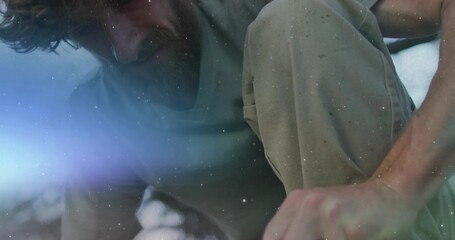 Crouching man wearing dusty cargo pants and grey shirt backlit outdoors, with bluish lens flare