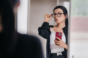 A confident woman captures her reflection while adjusting eyeglasses, exuding poise and charisma in...