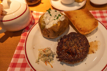A baked potato with sour cream, bread, and grilled meat on a dinner plate. Cozy restaurant meal served on checkered tablecloth.