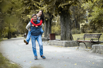 The playful couple enjoys a joyful piggyback ride in the park, displaying their love and happiness; a perfect moment captured amidst the beautiful autumn foliage and scenery.
