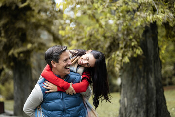 In the vibrant park, a couple shares a tender moment as the man carries the woman on his back, radiating joy and affection amidst the beautiful autumn scenery.