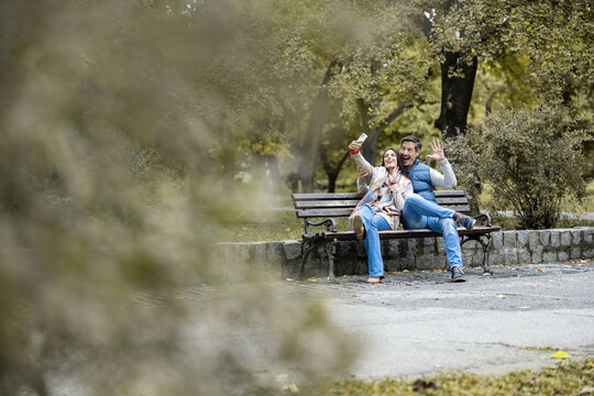 A happy couple enjoying a playful moment taking selfies while cozily seated on a bench in a park, capturing memories during a rainy day and highlighting their love and fun.