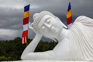 Closeup of the giant sleeping Buddha statue at the temple of Vihara Dharma Giri in northern Bali, Indonesia. Flags and cloudy sky in the background.
