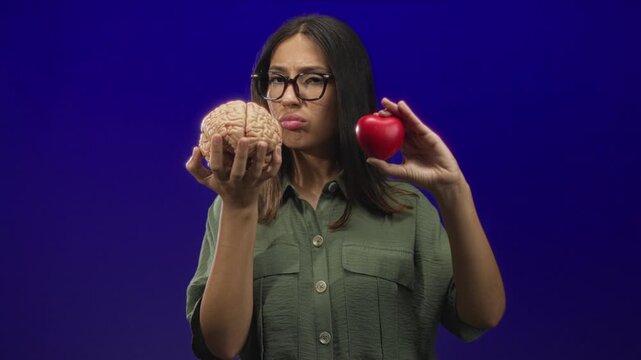Young hispanic brunette woman holding model brain and red heart in studio on blue purple backdrop; conflict decision.