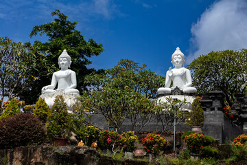 A pair of white Buddha statues adorn the garden of the Buddhist monastery Brama Vihara Aram in northern Bali, Indonesia. Blue sky and clouds in the background. 
