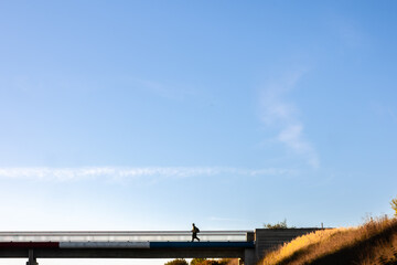 Fototapeta premium Person walks on bridge above highway. Silhouette under clear sky.