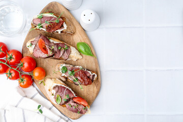 Delicious bruschettas with roast beef served on white tiled table, flat lay. Space for text