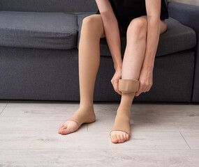 Female model wearing compression socks sitting on grey sofa indoors demonstrating healthy lifestyle, vein care and prevention of venous disease