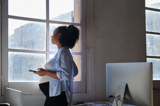 side view Pregnant black woman using phone standing by office window during a coffee break
