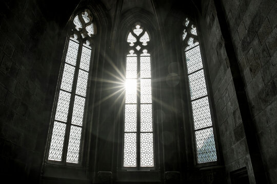 Black and white image of gothic arched stained glass windows inside Mont Saint Michel, with sunlight beaming through the glass and casting rays across the historic stone architecture
 - Powered by Adobe