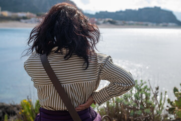 Woman photographed from behind while observing the panorama of the Tono bay, in Milazzo, along the Sicilian coast in Italy. The woman has long dark hair and wears a striped shirt