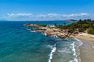 An aerial view of Rose Bay in Bowen, Queensland, reveals a stunning tropical coastline with turquoise waters, golden sand, and rocky headlands. The scene highlights coastal beauty and vibrant color.