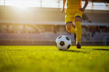 Soccer player in yellow uniform running with ball on green field during sunset, stadium in background, sports action