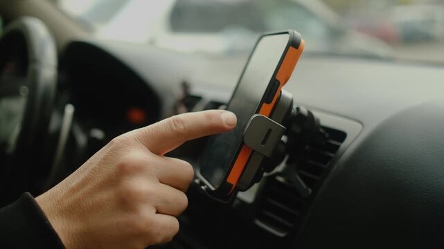 The driver's hand points to an orange smartphone mounted on the car's dashboard, indicating inattention. The driver is adjusting the car's navigation system.