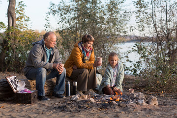 Little child girl with grandpa and grandmother Having a picnic on the beach on a sunny autumn day. Concept of friendly family.  Family  roasts marshmallows and sausages on the campfire.
