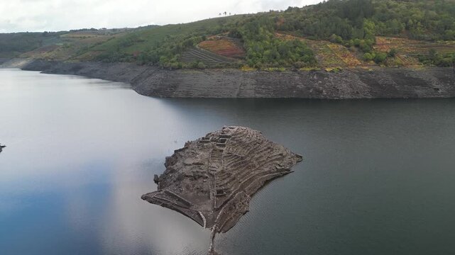 Aerial view of the ancient ruins of Castro Candaz rise majestically from the dark lake waters, a stark contrast against the green hills. Belesar, Spain - 02 November 2025