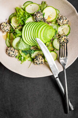 Fresh gourmet green salad with mixed greens, thinly sliced cucumber, and round goat cheese balls coated in black and white sesame seeds, served with a fork and knife
