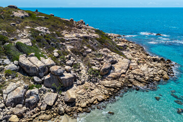 An aerial view of Rose Bay in Bowen, Queensland, reveals a stunning tropical coastline with turquoise waters, golden sand, and rocky headlands. The scene highlights coastal beauty and vibrant color.