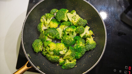 Fresh broccoli florets cooking in a non-stick pan on the stovetop. Vibrant green vegetables being sautéed with heat, showing signs of caramelization. Overhead shot with natural cooking process