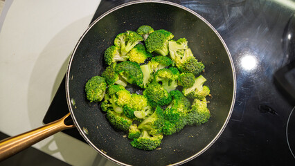 Fresh broccoli florets cooking in a non-stick pan on the stovetop. Vibrant green vegetables being sautéed with heat, showing signs of caramelization. Overhead shot with natural cooking process