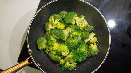 Fresh broccoli florets cooking in a non-stick pan on the stovetop. Vibrant green vegetables being sautéed with heat, showing signs of caramelization. Overhead shot with natural cooking process