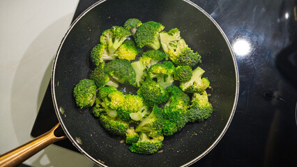 Fresh broccoli florets cooking in a non-stick pan on the stovetop. Vibrant green vegetables being sautéed with heat, showing signs of caramelization. Overhead shot with natural cooking process
