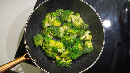 Fresh broccoli florets cooking in a non-stick pan on the stovetop. Vibrant green vegetables being sautéed with heat, showing signs of caramelization. Overhead shot with natural cooking process
