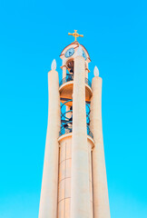 Modern bell tower of the Resurrection of Christ Orthodox Cathedral in Tirana, Albania. Its unique design features a clock face and a spiraling staircase, topped by golden cross against clear blue sky
