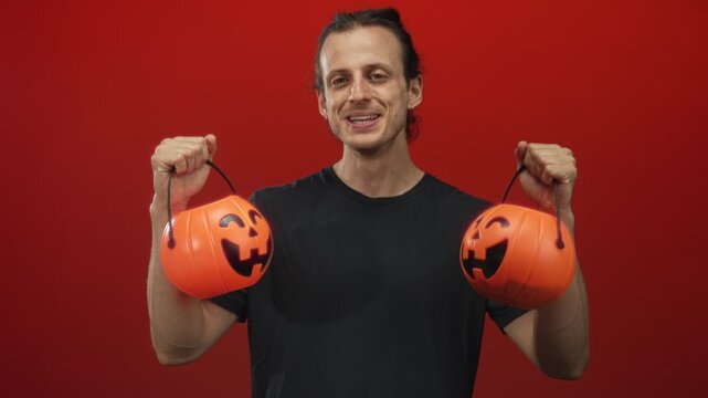Young hispanic man with long hair holding two smiling jack o lantern pumpkin buckets by hands in a red studio; playful halloween fun.