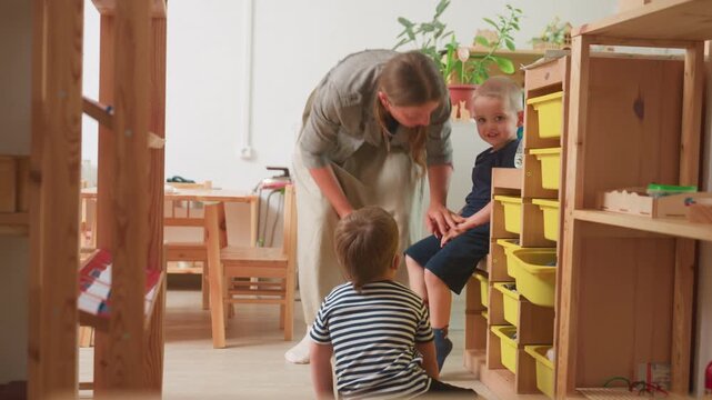 caregiver kneels beside boy in blue by classroom storage bins, guiding hand and giving reassurance, while second child in striped top watches from floor as they go