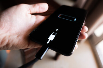 A black smartphone with a low battery icon is connected to a charger on a wooden surface. The charger is plugged into a power outlet.
