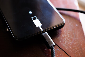 A black smartphone with a low battery icon is connected to a charger on a wooden surface. The charger is plugged into a power outlet.