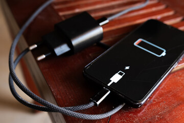 A black smartphone with a low battery icon is connected to a charger on a wooden surface. The charger is plugged into a power outlet.