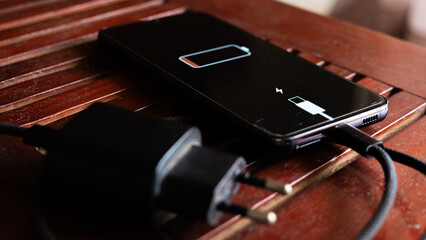 A black smartphone with a low battery icon is connected to a charger on a wooden surface. The charger is plugged into a power outlet.