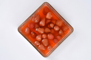 A square glass dish is filled with strawberry compote featuring whole and halved strawberries in a light orange-pink syrup, photographed from above on a neutral white background