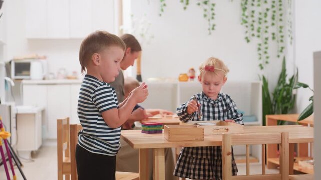 boy with hearing aid stacks colorful tiles at wooden table during preschool session, teacher assists in background while blonde girl in plaid dress works with tray, inclusive classroom promotes focus - Powered by Adobe