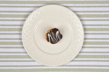 A single, chocolate-glazed profiterole with white drizzle rests on a white decorative plate placed centrally on a green and white striped tablecloth