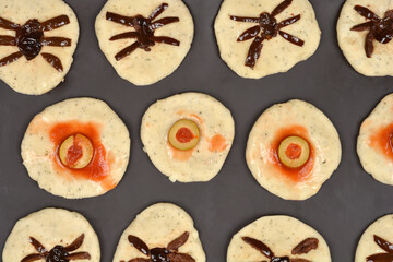 An overhead shot of unbaked Halloween appetizers made from raw dough, topped with olive slices and tomato sauce for an eyeball or spider effect