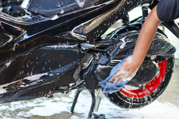 Person washing a glossy black scooter body and rear wheel, covered in thick white soap suds, highlighting vehicle maintenance and care process. © Jumain