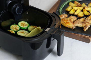 Air fryer with grilled vegetables, chicken wings, potato wedges and spices on white marble table, closeup