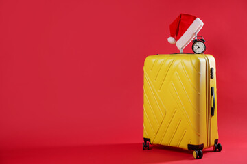 Christmas vacation. Yellow suitcase with Santa hat and alarm clock on red background, space for text