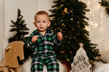 A little boy sits in a checkered green pajamas near green Christmas trees