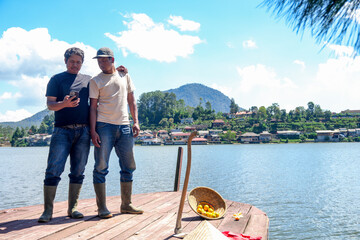 Two Farmers Checking Their Phone on Peaceful Lake Pier Overlooking Village on Beautiful Sunny Day