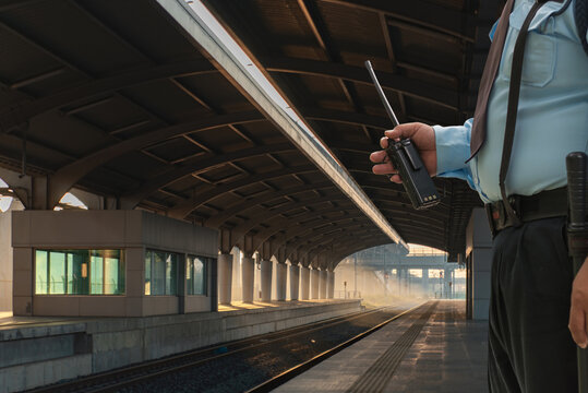 A uniformed security guard holds a walkie-talkie at a quiet, empty train station platform under morning sunlight, ensuring passenger safety. - Powered by Adobe