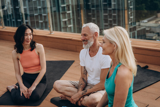 Senior man with grey beard sits on rooftop deck listening and smiling in break during yoga session