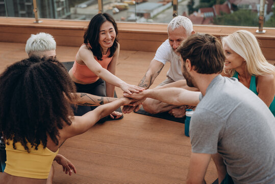 Group of happy people join hands in symbolic gesture expressing happiness after outdoor yoga class on hotel rooftop
