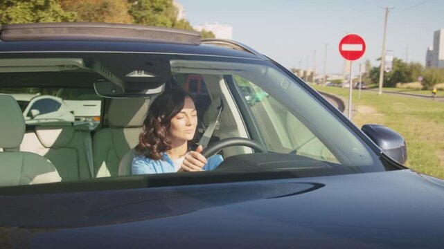 Pretty Caucasian female with wavy brown hair steering car while making turn on city street. Woman smiling and enjoying smooth drive in daylight. Buildings reflecting on shiny vehicle surface.